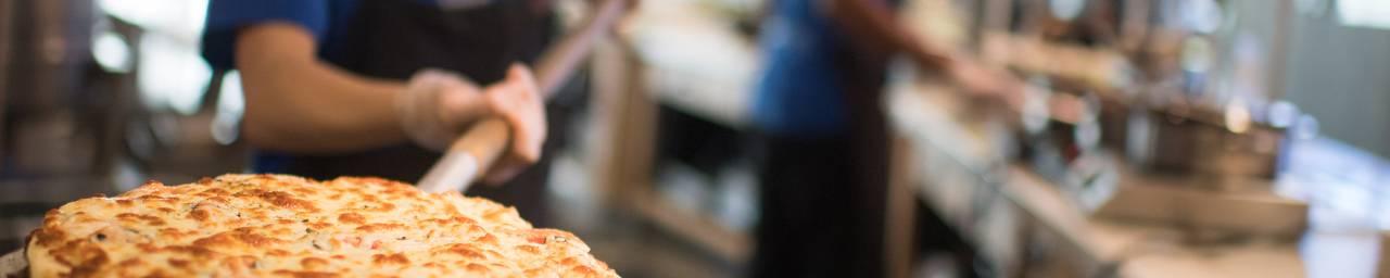一个刚从烤箱里出来的芝士披萨的特写，由一个校园食堂的学生员工拿着